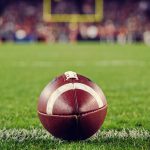 A closeup of an American football on an American football field, with the goalpost blurred in the far background