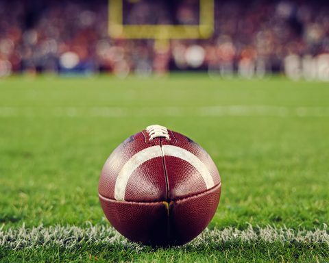 A closeup of an American football on an American football field, with the goalpost blurred in the far background